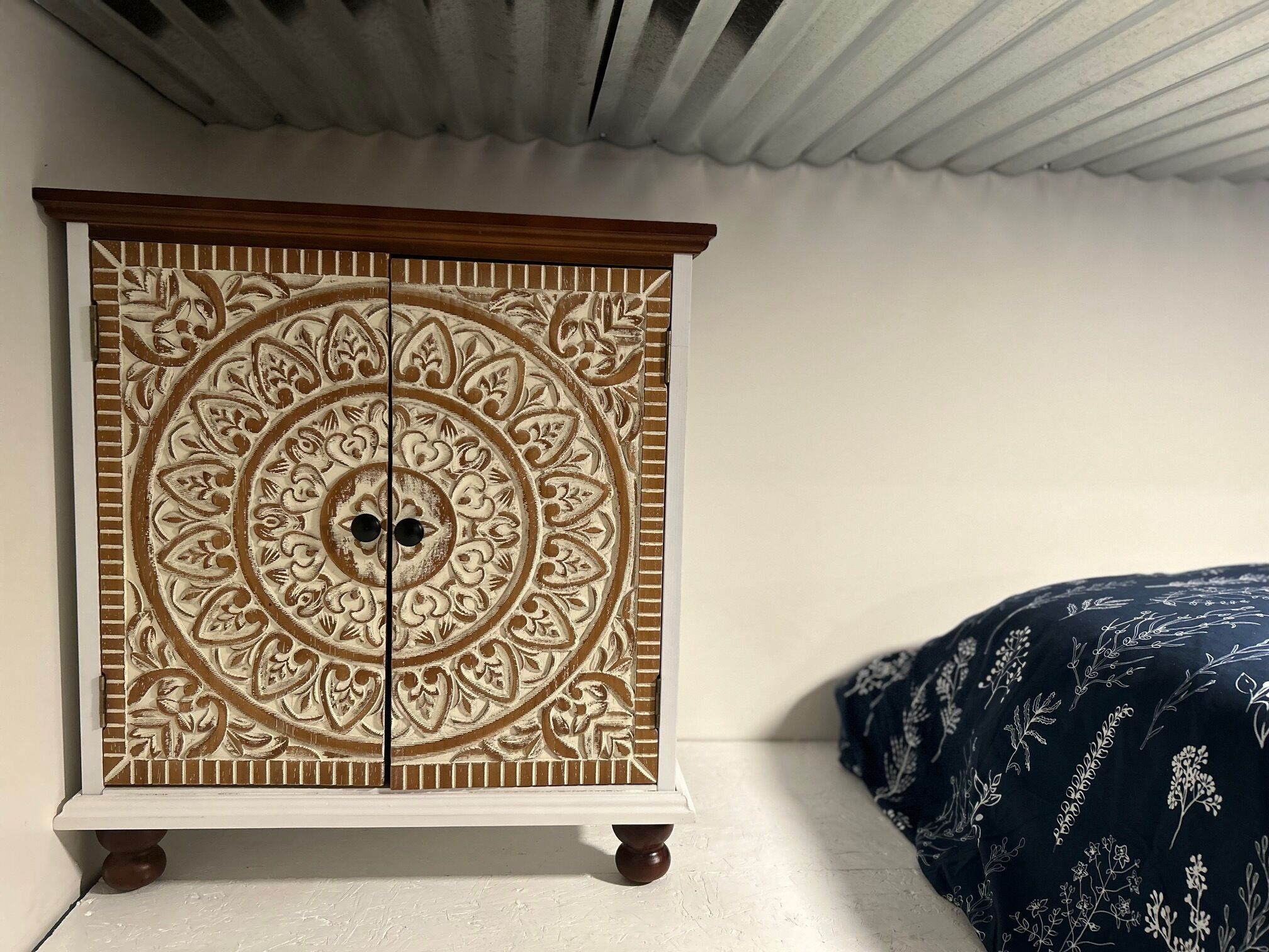 Un petit meuble d’apparat, richement décoré de motifs complexes, placé à côté d’un lit recouvert d’une couverture à motifs sombres.