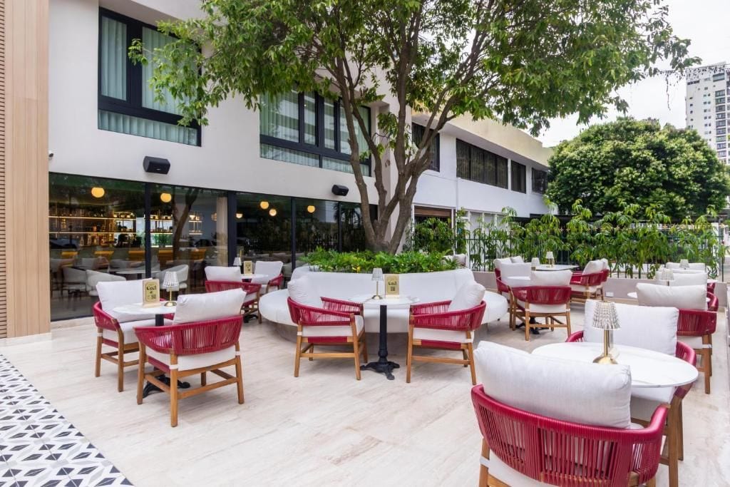 An outdoor seating area with wicker chairs and tables, a tree, and a building with large windows in the background.