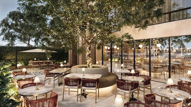 An outdoor seating area with round tables and wicker chairs, surrounded by trees and a large window overlooking the city. - Berthas Collection By EM Hotels (Cartagena)
