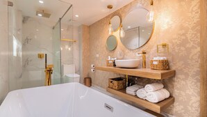 A modern bathroom with a large white bathtub, glass shower enclosure, and wooden shelves with toiletries. - Berthas Collection By EM Hotels (Cartagena)