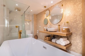 A modern bathroom with a large white bathtub, glass shower enclosure, and wooden shelves with toiletries. - Berthas Collection By EM Hotels (Cartagena)