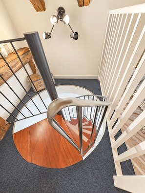 A spiral staircase with a wooden finish and metal railings, leading to a landing with a ceiling light fixture.