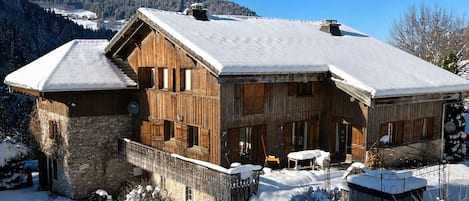A wooden chalet with a stone foundation, large windows, and a snow-covered roof, surrounded by a snowy landscape and mountains.