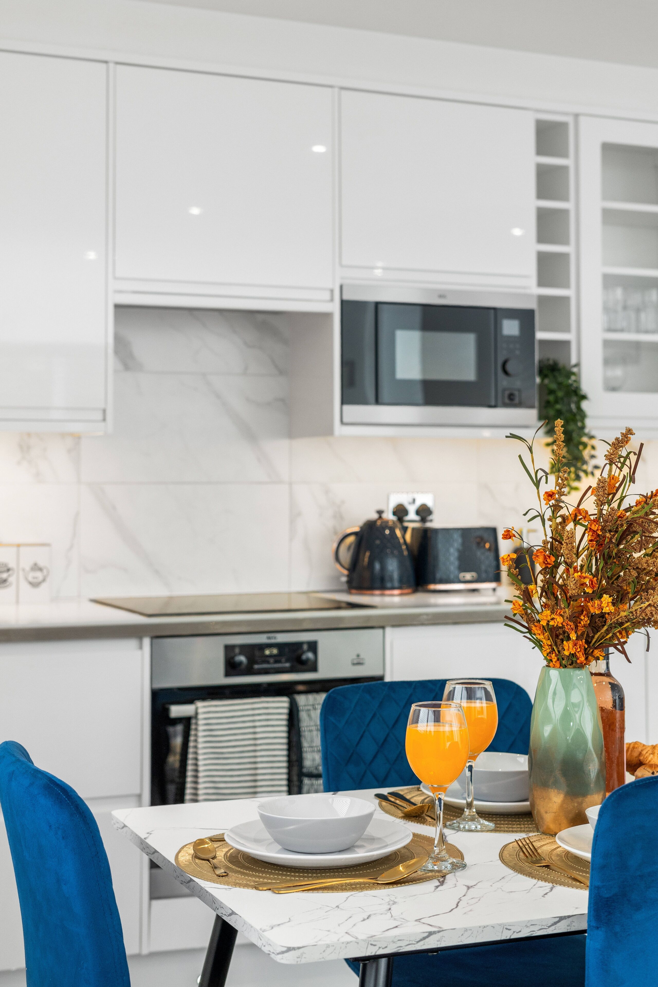 A modern kitchen with white cabinets, a microwave, and a dining table set with a vase of flowers, two glasses of orange juice, and a coffee maker.