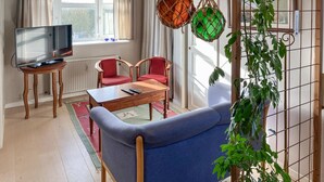 Ein Wohnzimmer mit Fernseher, zwei roten Sesseln, einem blauen Sofa, einem Holztisch und hängenden Wandteppichen aus Makramee.