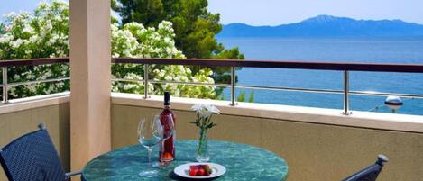 A balcony with a table set for two, featuring a bottle of red wine, a glass, and a plate with fruit. The balcony overlooks a body of water and a mountainous landscape.
