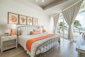 Ein Schlafzimmer mit Bett, Nachttischen, einem Fenster mit Blick auf den Strand und gerahmten Bildern an der Wand.