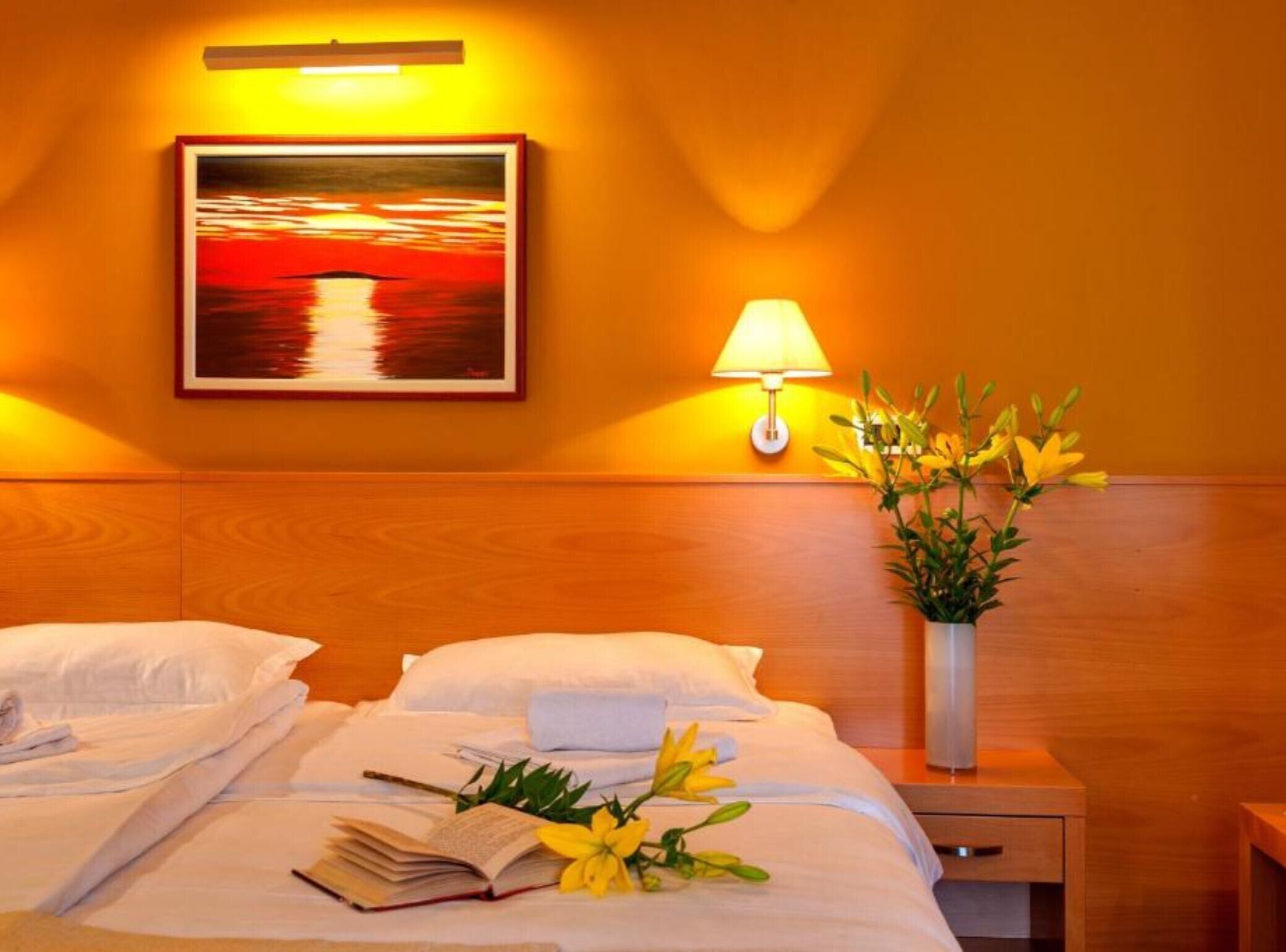 A hotel room with a bed, a bedside table with a lamp, a framed painting on the wall, and a vase with flowers on the nightstand.