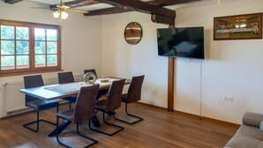Un comedor con una mesa de madera, seis sillas, un ventilador de techo, un reloj, un televisor y una ventana con vistas a árboles.