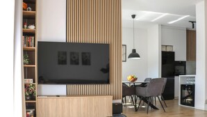 Een moderne woonkamer met een flatscreen televisie, een wand van houten panelen, een eethoek met stoelen en een keuken op de achtergrond.
