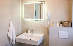 Une salle de bain moderne avec un lavabo blanc, un miroir doté d’un éclairage intégré, un sèche-cheveux et une petite plante en pot posée sur le meuble-lavabo.