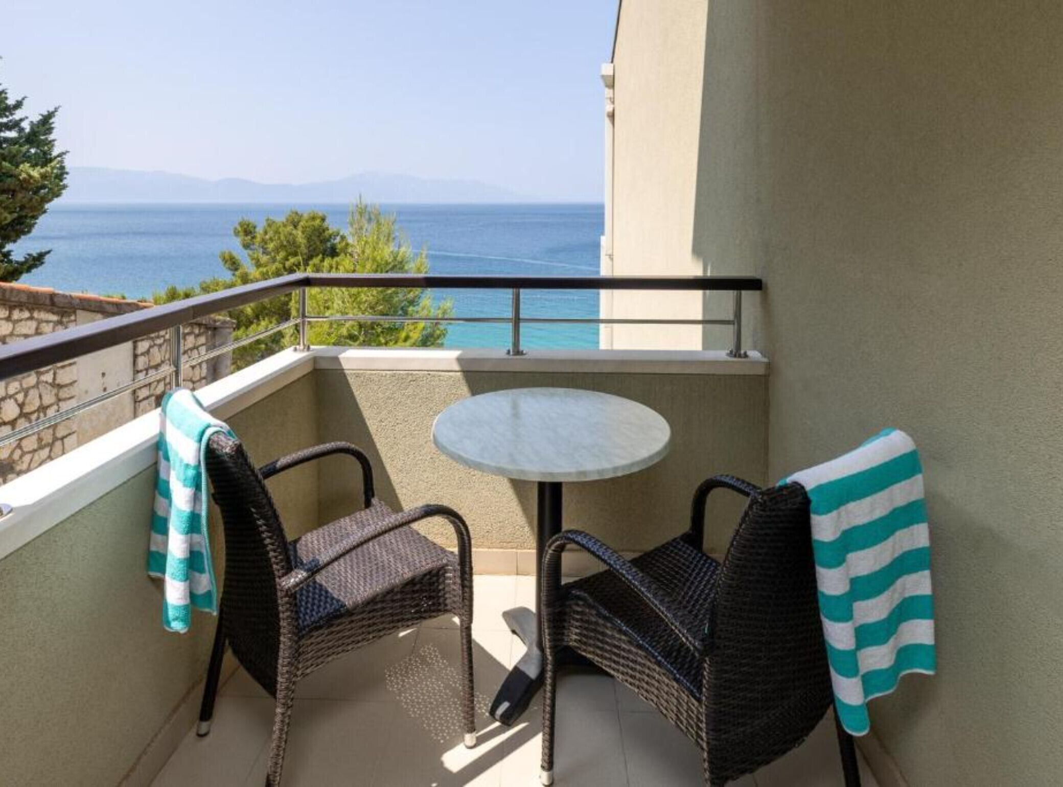 A balcony with two wicker chairs, a small round table, and a view of the sea and mountains.