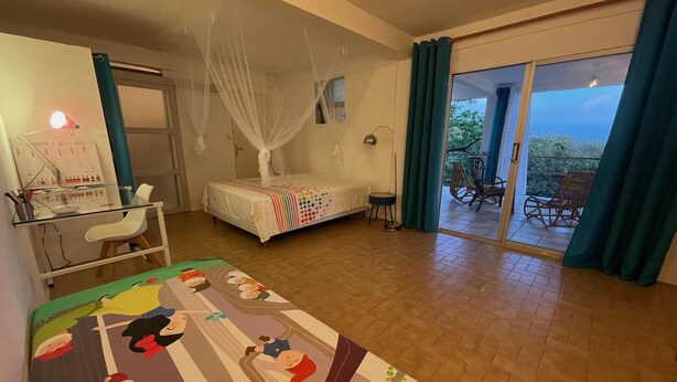 A bedroom with a bed, a desk, a chair, a small table, and a balcony with chairs. - Appartement vue mer - Hauteurs du Carbet - Kaz des Alizés Au frais - cats allowed (Le Carbet)