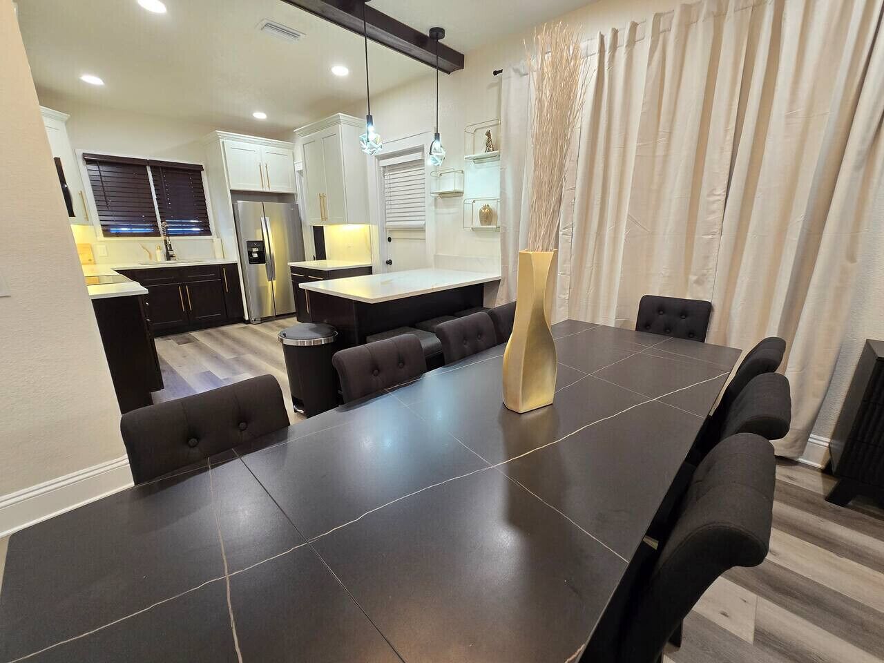 A modern dining area with a large table, cushioned seating, and a kitchen in the background.