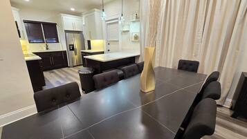 A modern dining area with a large table, cushioned seating, and a kitchen in the background.