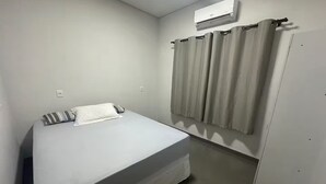 Un llit individual amb matalàs blanc i almoina amb ratlles, una finestra amb cortines beix i un aparell d'aire condicionat a la paret.