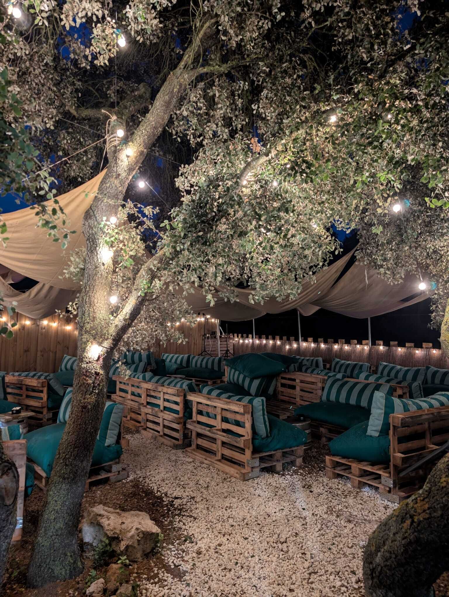 Un espacio de estar al aire libre con bancos de madera y cojines, rodeado por una pérgola y guirnaldas de luces.