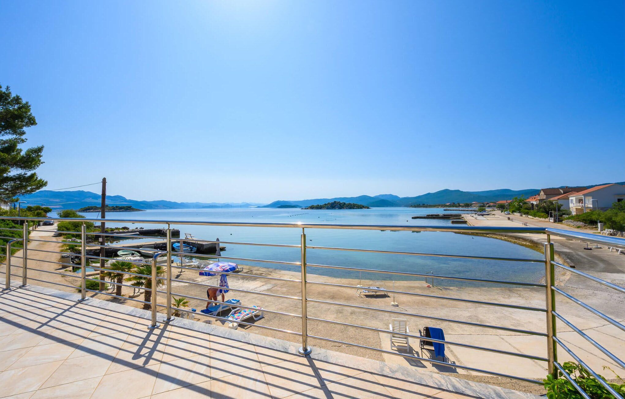 Une terrasse en bord de mer offrant une vue sur la mer et les îles au loin, une plage de sable et un petit espace aménagé en quai.