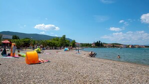 Une plage de galets où des gens se prélassent au soleil et se baignent, un ciel bleu dégagé et des bâtiments en arrière-plan.