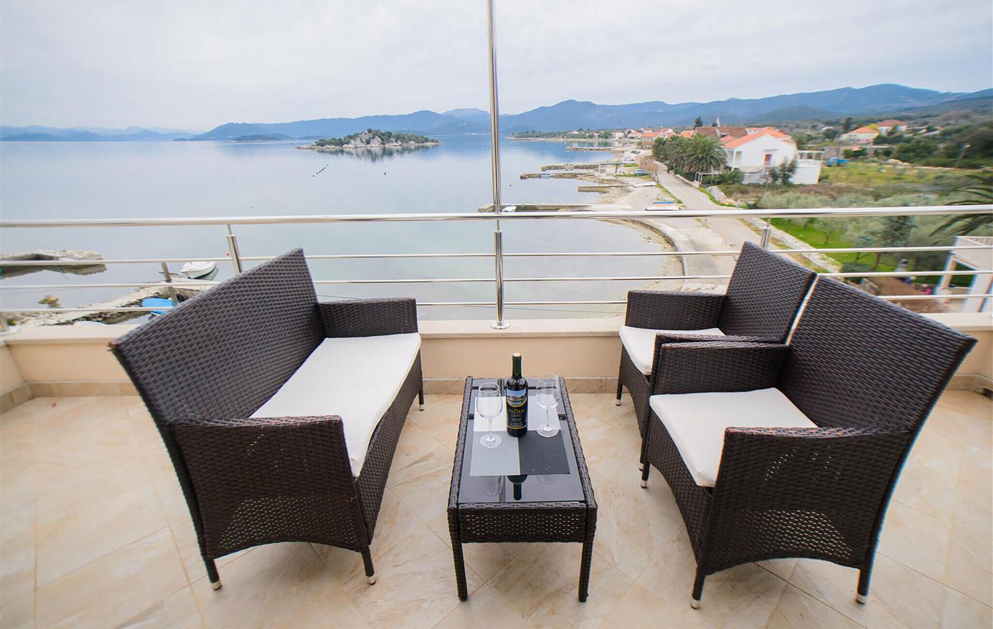 A balcony with wicker furniture and a table, overlooking a body of water and distant hills.