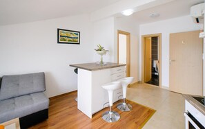 Een compacte leefruimte met een kitchenette, een grijze bank, een klein barretje en een houten vloer.