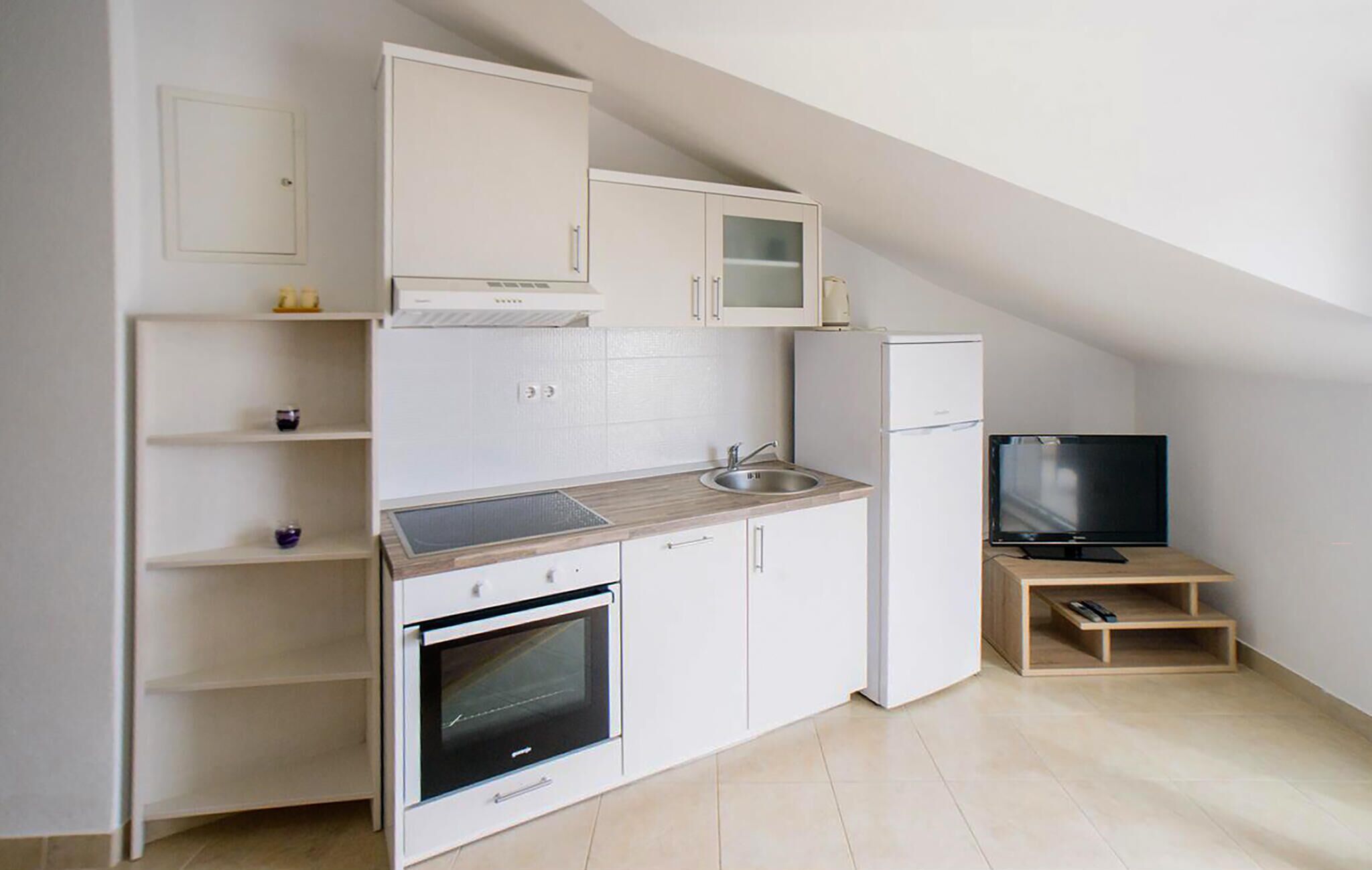 Een kleine keuken met witte kastjes, een ingebouwde oven, een koelkast en een televisie op een houten plank.