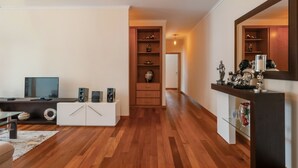 A modern living room with a wooden floor, a white entertainment unit, a television, and a decorative shelf with various items. - Freitas House by Homie (Canhas)