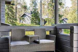 Et overdækket udendørs opholdsområde med fletmøbler og udsigt til huse i et skovområde.