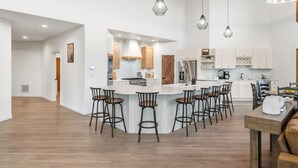 A modern kitchen with a central island, bar stools, pendant lighting, and a dining area with a wooden table.