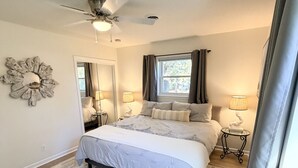 Ein Schlafzimmer mit Bett, Deckenventilator, Fenster mit Vorhängen, Spiegel und einem kleinen Tisch mit Lampe.
