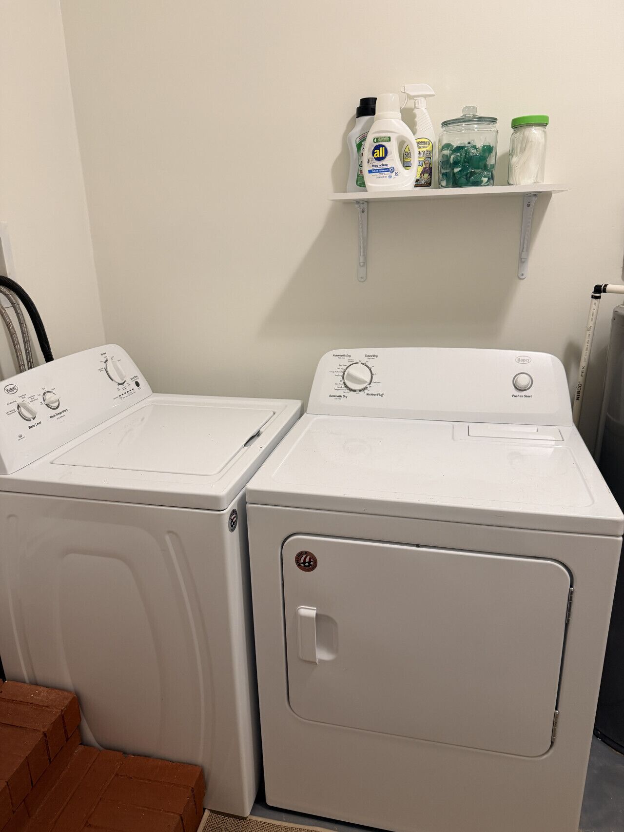 A laundry area with two front-loading washing machines, a shelf with cleaning supplies, and a brick wall.
