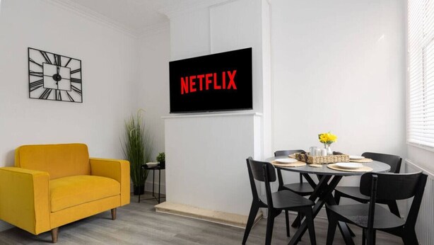 A modern living room with a yellow sofa, a dining table with black chairs, a black wall-mounted TV displaying "NETFLIX", and a decorative clock. - Charming Central Leicester 4 Bed Accommodation (Leicester)