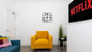 A room with a yellow armchair, a blue sofa, a wall-mounted Netflix sign, and a clock. - Charming Central Leicester 4 Bed Accommodation (Leicester)