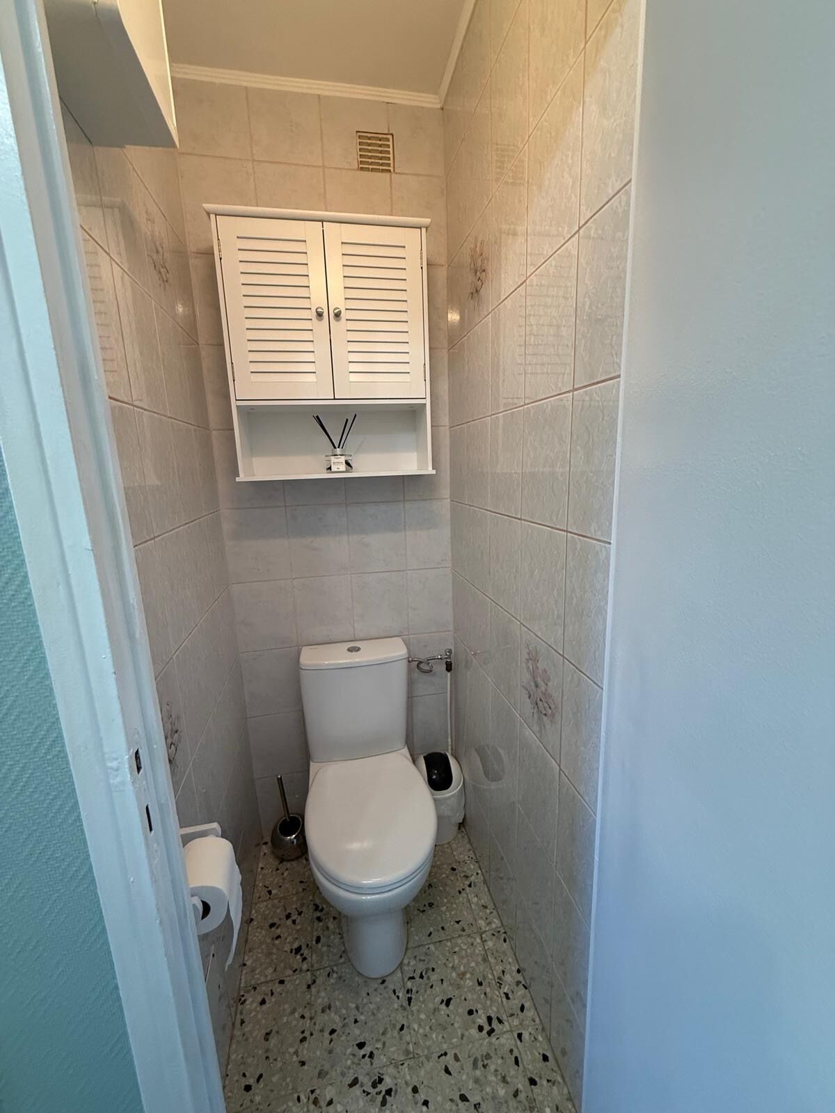 A small, narrow bathroom with a toilet, a white cabinet above it, and a trash bin to the side.