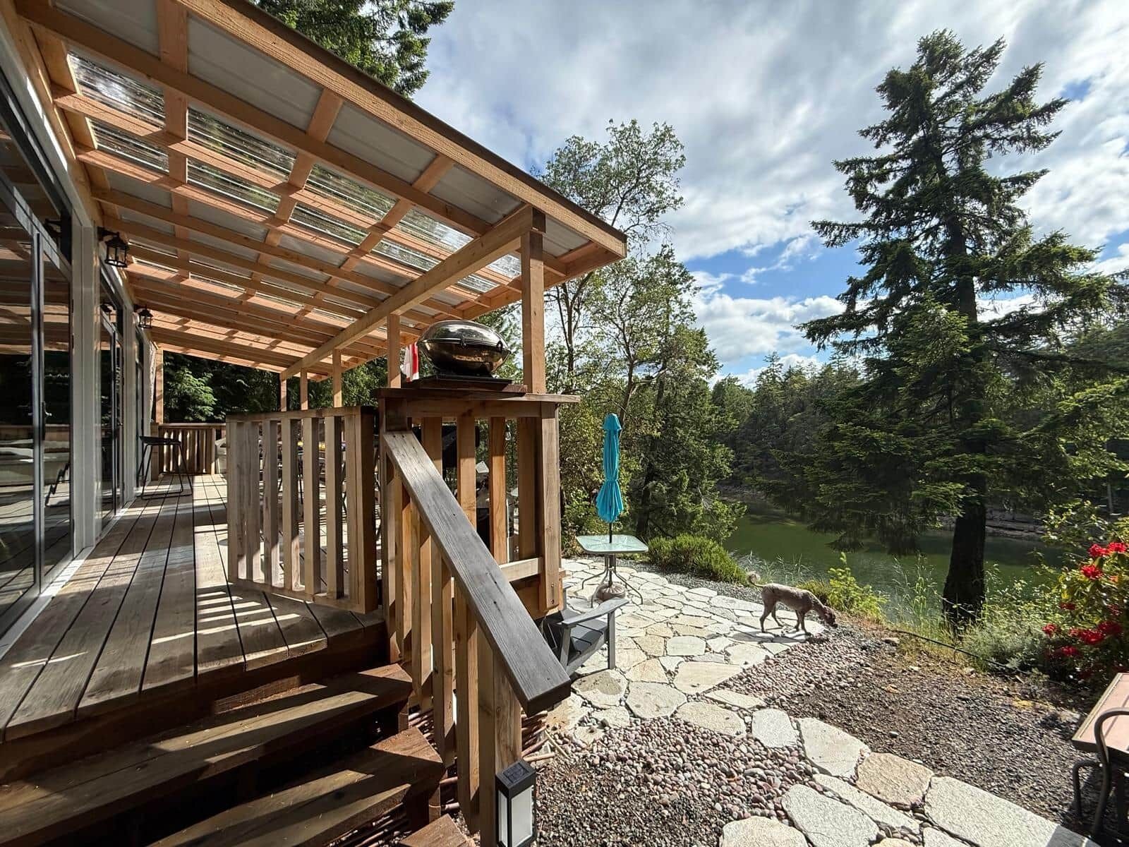 Une terrasse en bois avec une pergola, un patio en pierre et une vue sur un lac, avec un chien à proximité.