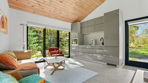 Ein modernes Wohnzimmer mit einer Couch, einem Couchtisch und einem roten Sessel. Man sieht eine Küche mit grauen Schränken und einem großen Fenster.