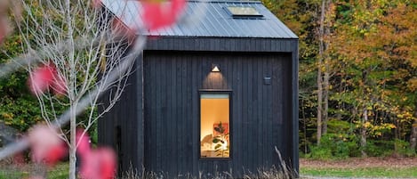 A small, dark wooden cabin with a visible interior window, surrounded by trees and grass.