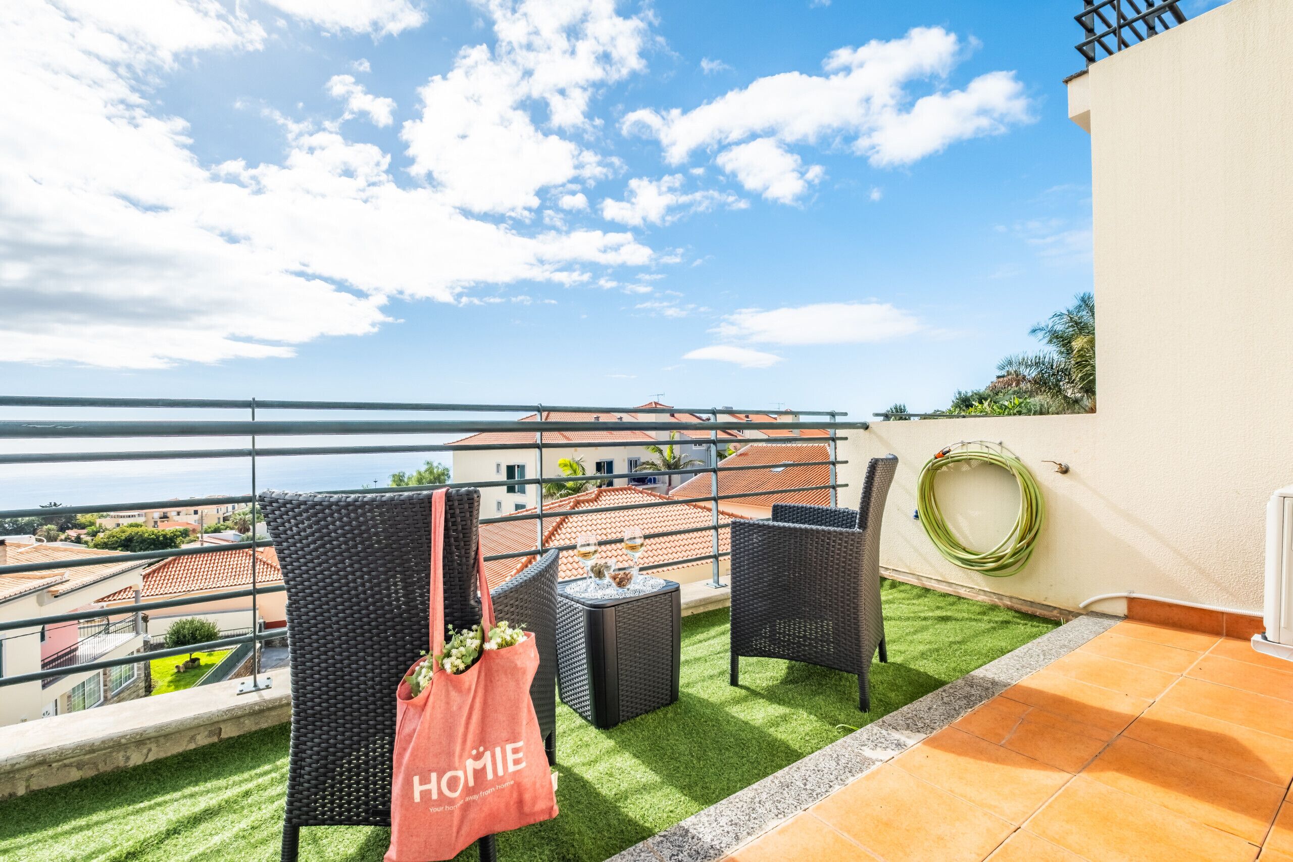 A balcony with wicker furniture, a table with glasses, and a view of the sea and buildings.