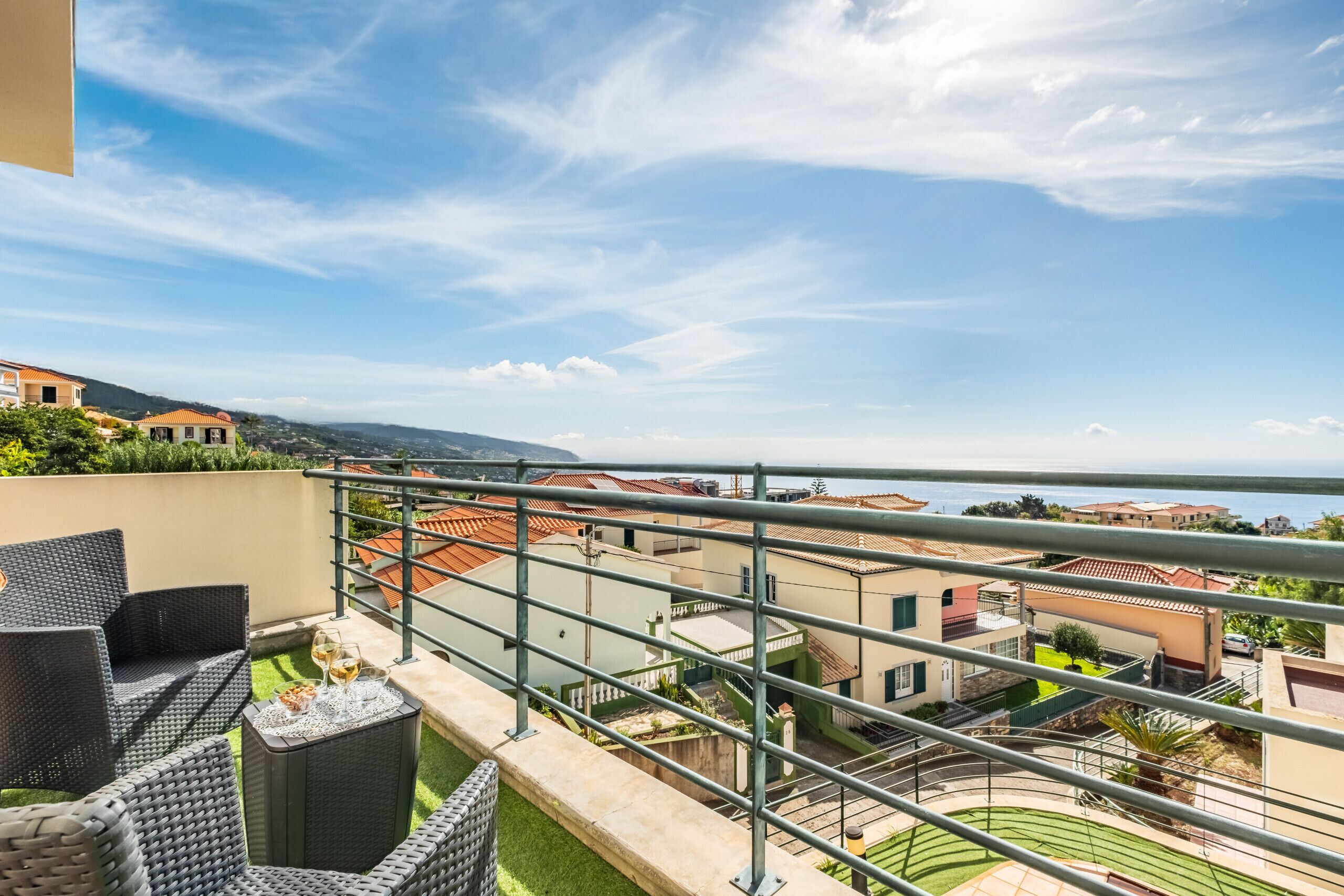 A balcony with outdoor furniture, a view of the sea, and a residential area with houses.
