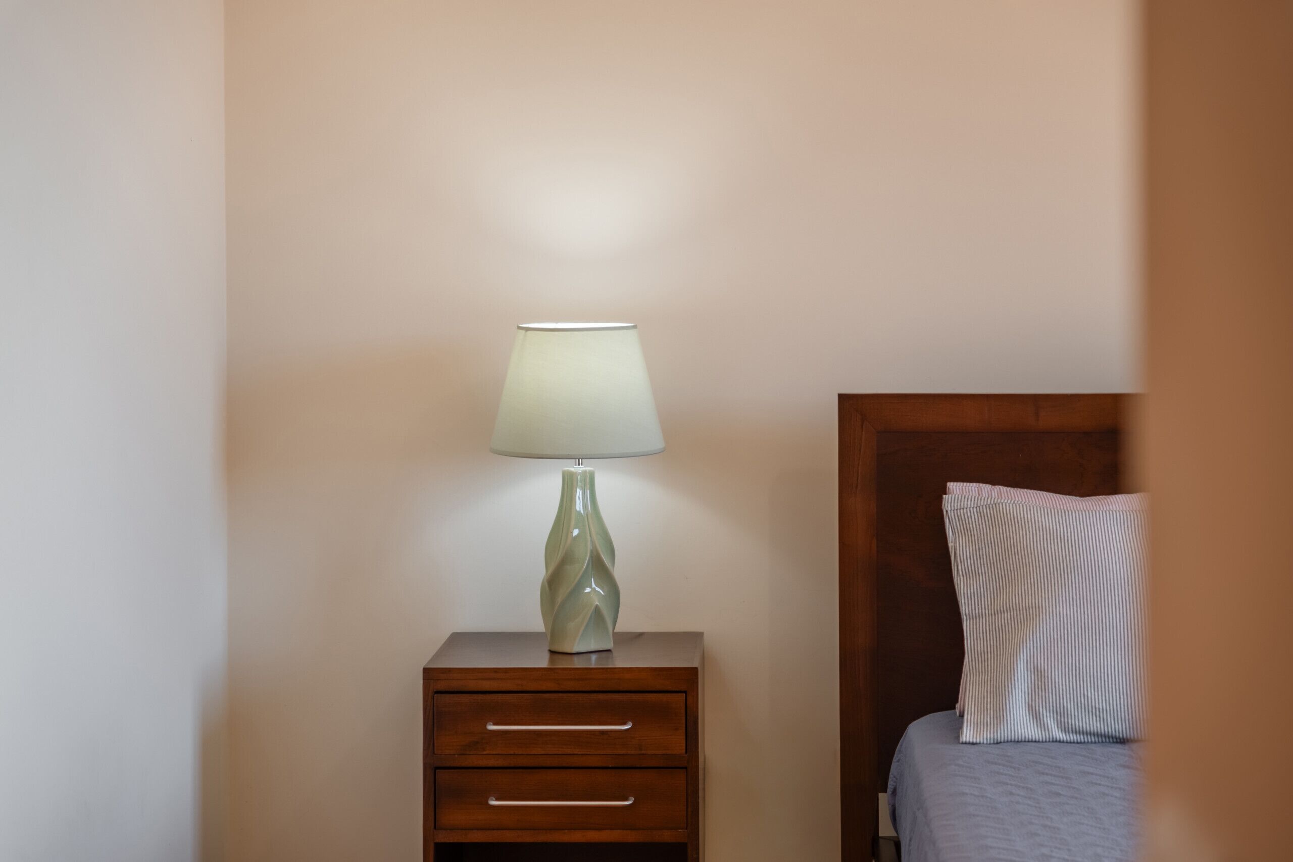 Et sengebord med lampe, en skuffe og en seng med pude.