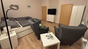 Un espace de vie compact comprenant un canapé-lit, une petite table avec une bouteille et des verres, un ventilateur de plafond et un téléviseur posé sur une commode.