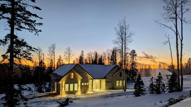Lumipeitteinen maisema, jossa on kivitalo, jossa on harjakatto ja suuret ikkunat. Taloa ympäröivät korkeat puut ja taivaalla on kirkasta.