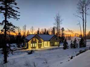 Un paysage enneigé avec une maison en pierre dotée d’un toit à pignon et de grandes fenêtres, entourée de grands arbres et surplombée d’un ciel dégagé.