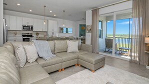 A modern living room with a sectional sofa, a dining area with chairs, a kitchen with white cabinets and appliances, and a balcony with a view. - Top-Floor Babcock Ranch Condo Near Fort Myers • Pool, Golf, SW Florida (Punta Gorda)