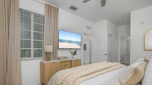 A bedroom with a large flat-screen TV, a bed with white bedding, a wooden nightstand, and a window with beige curtains. - Top-Floor Babcock Ranch Condo Near Fort Myers • Pool, Golf, SW Florida (Punta Gorda)