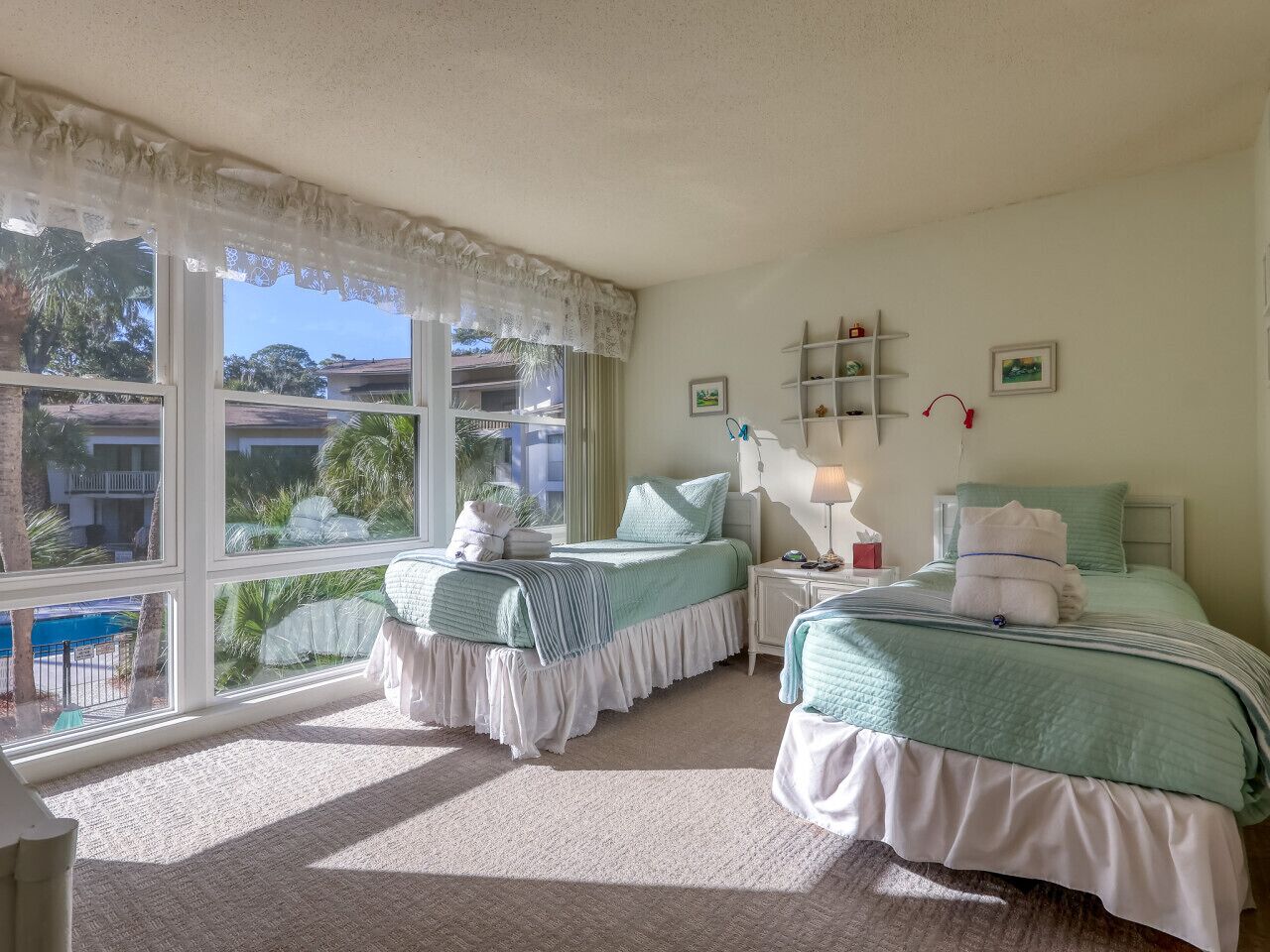 A bedroom with two beds, large windows, a view of a pool, and a small shelf with decorative items.