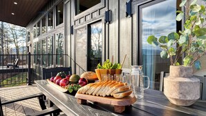 Una mesa de madera con diversas frutas, verduras y pan, una jarra de vidrio y una planta en maceta en un balcón con vistas a árboles y un edificio.