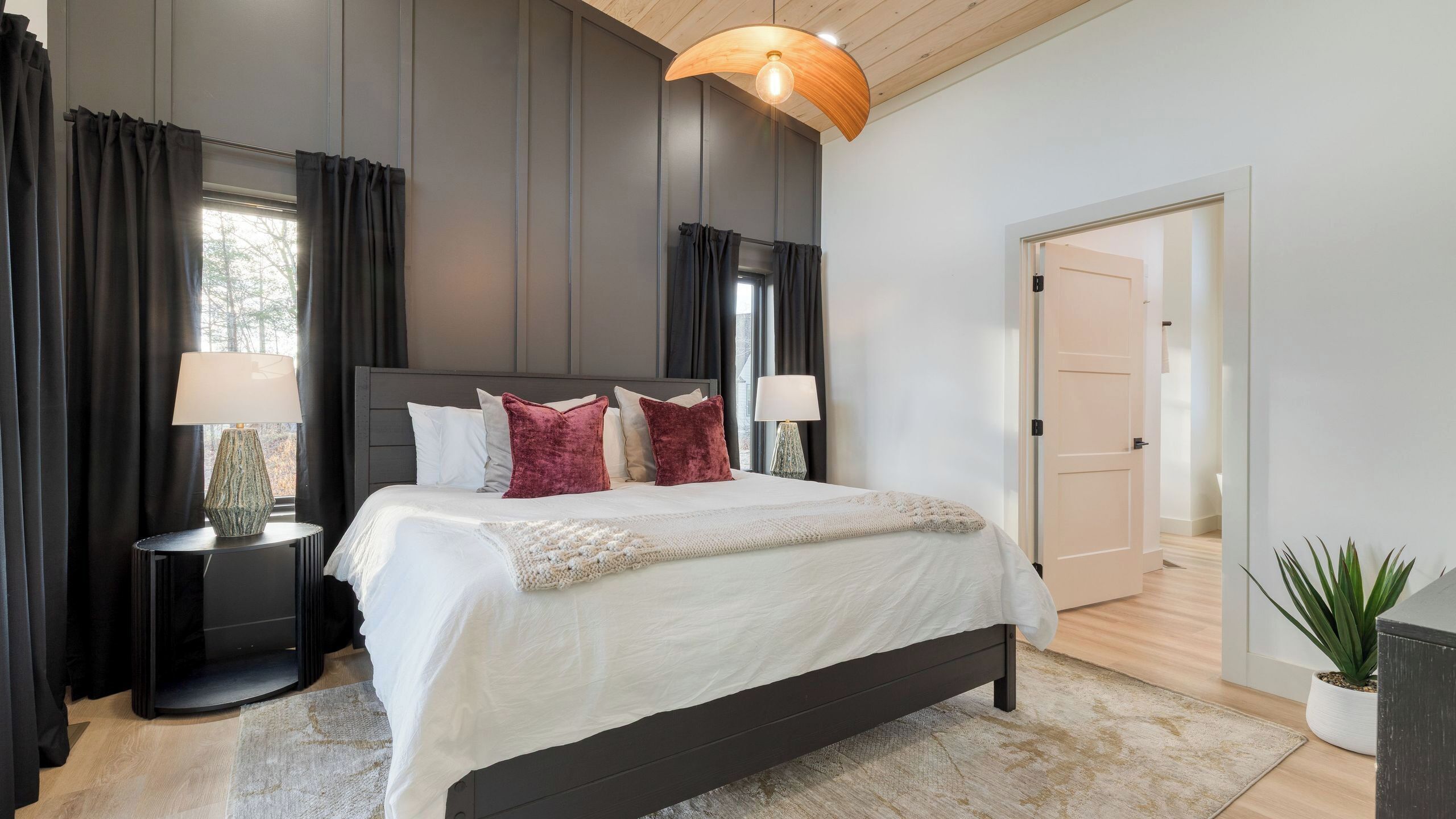 A modern bedroom with a large bed, dark wood headboard, two bedside tables with lamps, and a large window with curtains.