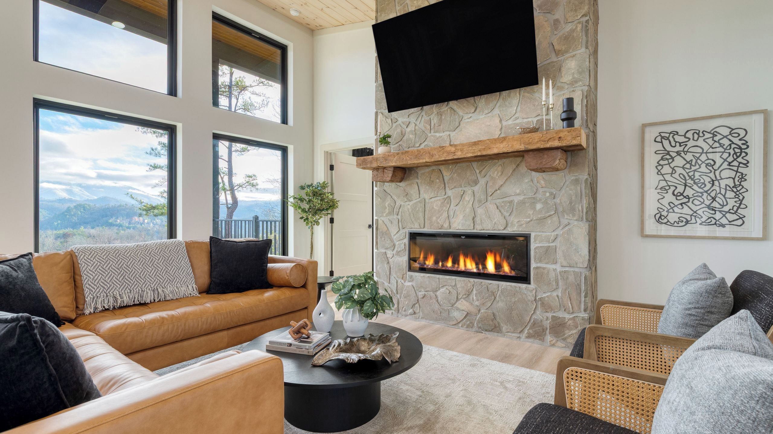 A modern living room with a stone fireplace, large windows, and a flat-screen TV mounted on the wall.
