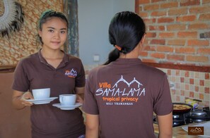 Deux personnes portant des chemises marron, avec l’inscription « Villa Samaluma, intimité tropicale » imprimée dans le dos, servent du café dans un restaurant dont le décor comprend un mur de briques et un mur arrière recouvert de carreaux.